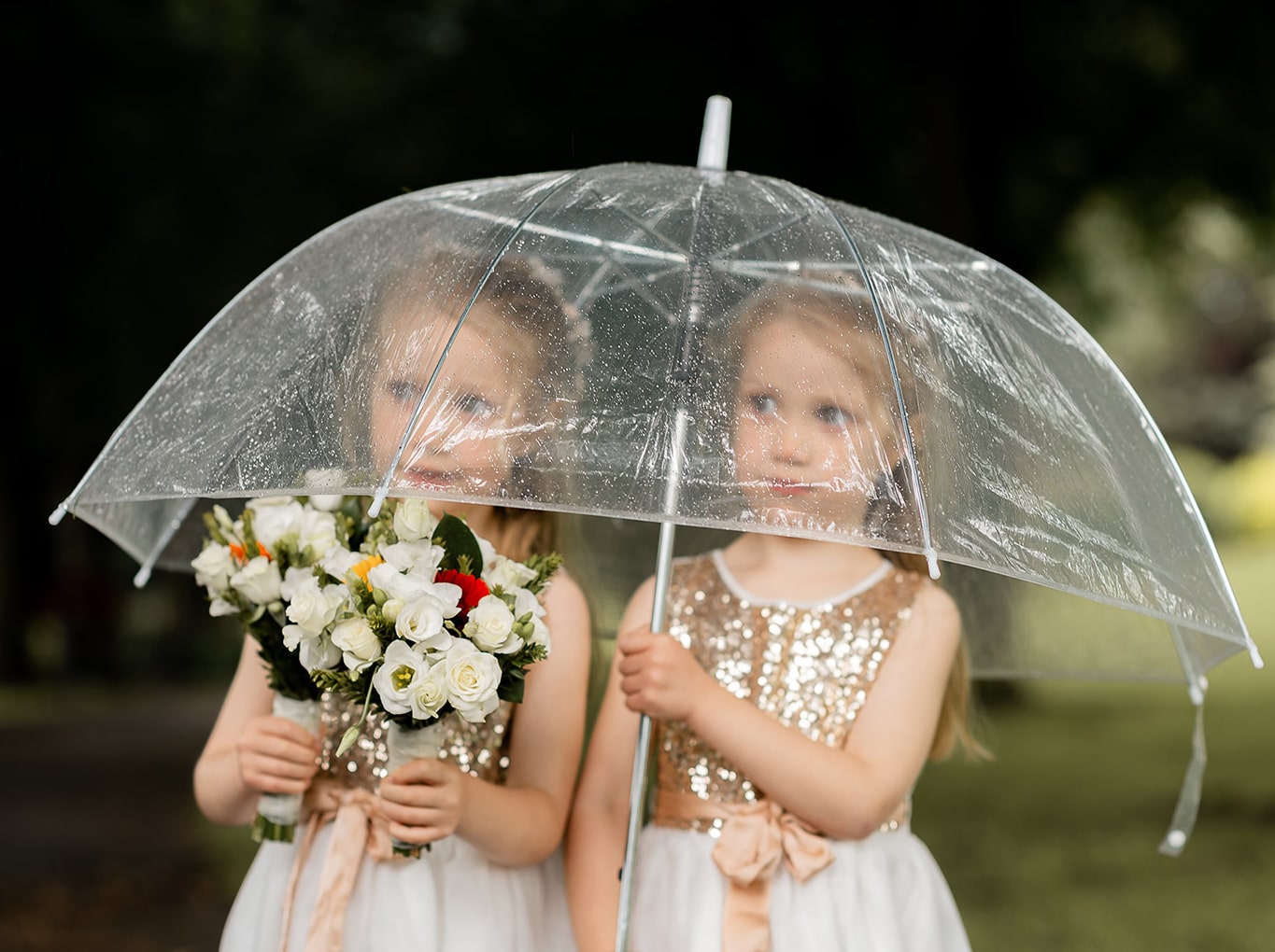 Cute photo of twin girl bridesmaids sharing a transparent umbrella in the rain, holding a bouquet of flowers